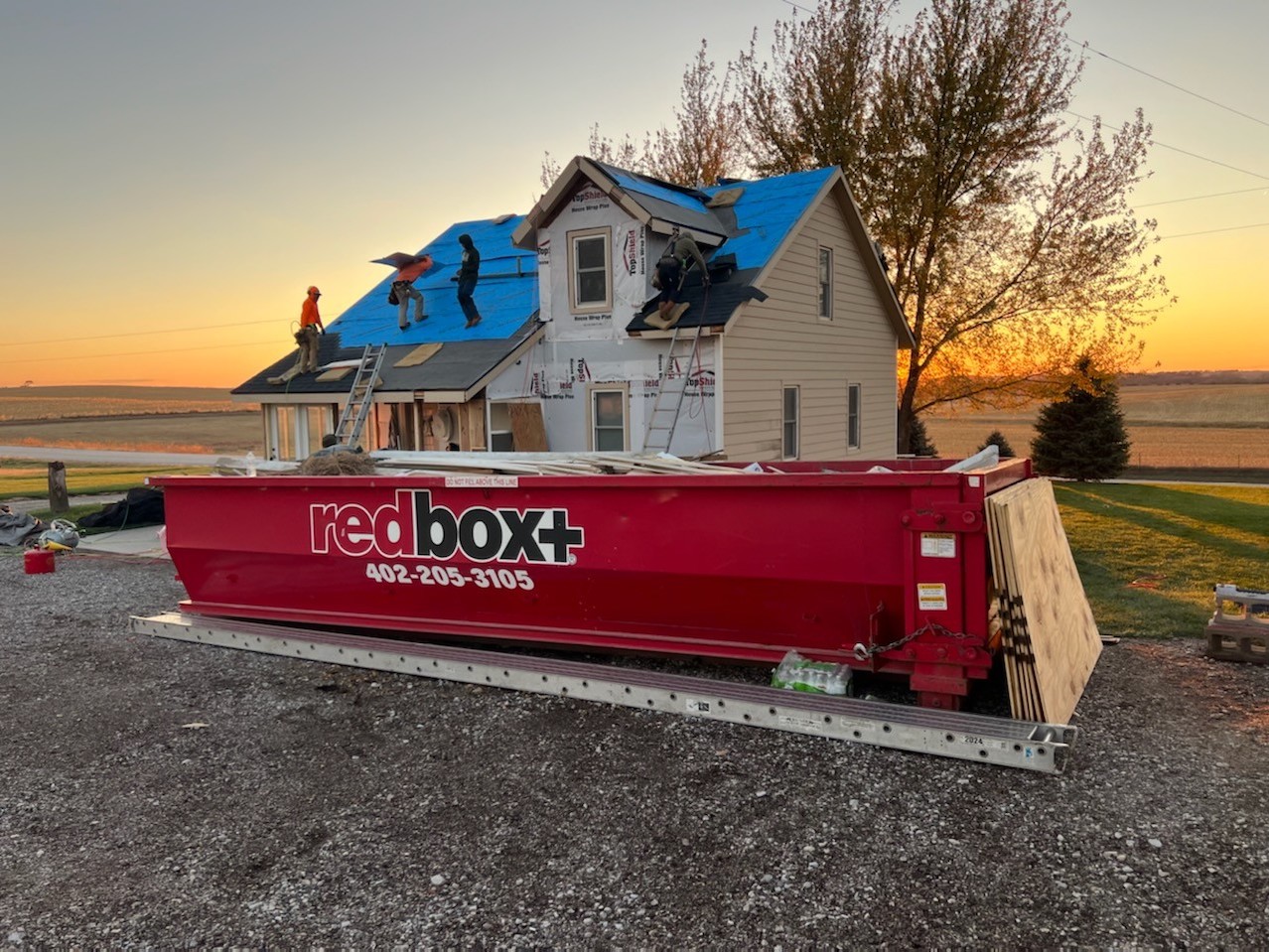 Red redbox+ dumpster in front of a storm-damaged house at sunset